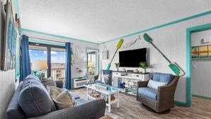 Un salon avec un canapé, une table basse et un fauteuil en osier. Il y a un téléviseur accroché au mur et une fenêtre avec des rideaux.