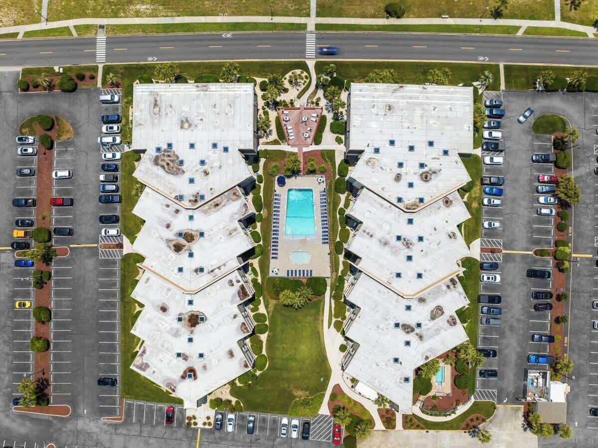 An aerial view of a hotel with a central swimming pool, multiple parking lots, and landscaped areas.