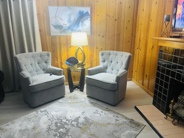 A living room with two grey armchairs, a glass coffee table, a modern abstract painting, a floor lamp, and a tiled fireplace. - Historic home on West Main (Paragould)