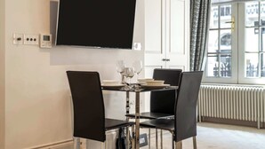 A modern dining area with a large flat-screen TV mounted on the wall, a dining table set with plates and wine glasses, and a window with a view of buildings. - Modern Studio in Mayfair next to Hyde Park (London)