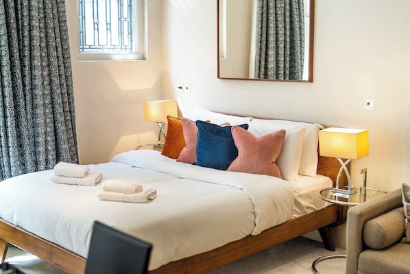 A neatly made bed with white linens and pillows, two bedside lamps, a mirror above the bed, and a window with curtains. - Modern Studio in Mayfair next to Hyde Park (London)
