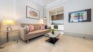 A modern living room with a sofa, a coffee table, and a lamp. There is a kitchen visible in the background. - Modern Studio in Mayfair next to Hyde Park (London)