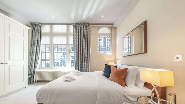 A hotel room with a large bed, a bedside lamp, a window with curtains, and a radiator. - Modern Studio in Mayfair next to Hyde Park (London)