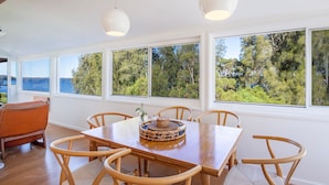 A dining area with a wooden table and chairs, large windows with a view of trees and the sea, and a hanging light fixture.