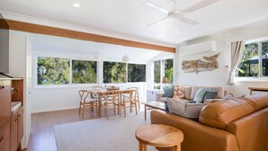 A spacious living area with a dining table, sofa, and large windows offering a view of greenery.