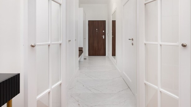 A hallway with white doors, a wooden door at the end, and a marble floor.