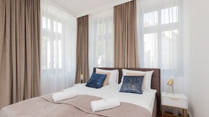 A neatly made bed with a beige quilt, two blue pillows, and a white pillow. There is a white bedside table with a lamp, and the room has large windows with sheer curtains.