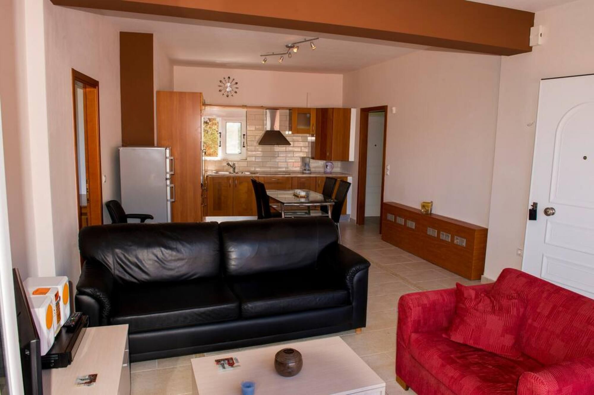 A living room with a black leather sofa, a red sofa, and a coffee table with a vase. There is a kitchen in the background with wooden cabinets and a dining area.