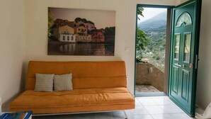 A room with a sofa, a painting of a coastal scene, and a door leading to a balcony with a view of a mountainous landscape. - Myrtos Cottages Polimnia (Kefalonia)