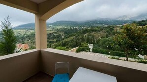 A balcony with a table and a chair, overlooking a mountainous landscape with greenery and distant buildings. - Myrtos Cottages Polimnia (Kefalonia)