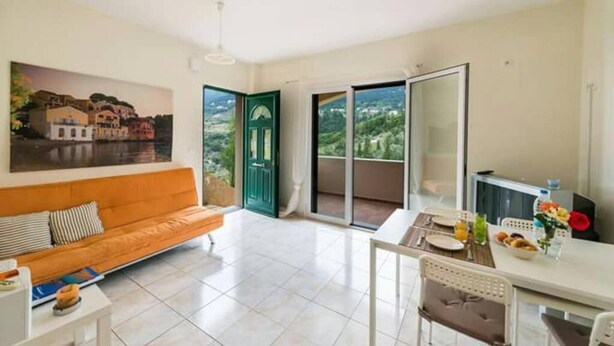 A bright living room with a sofa, a dining table with chairs, a TV, and a view of a green door and a balcony with a cityscape. - Myrtos Cottages Polimnia (Kefalonia)