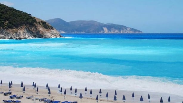 Ein Strand mit klarem, blauem Wasser, einem Sandufer und einer hügeligen Landschaft im Hintergrund.