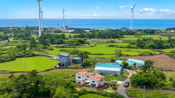 A rural landscape with wind turbines, green fields, and a house with a red roof. - Jeju West Dokchae Pension (Jeju City)
