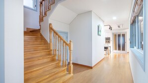 A wooden staircase leading to a landing with a door, a kitchen area with cabinets and a countertop, and a window with blinds. - Jeju West Dokchae Pension (Jeju City)