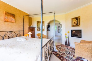 A four-poster bed with a white bedspread, a wicker chair, a patterned rug, and a painting on the wall.