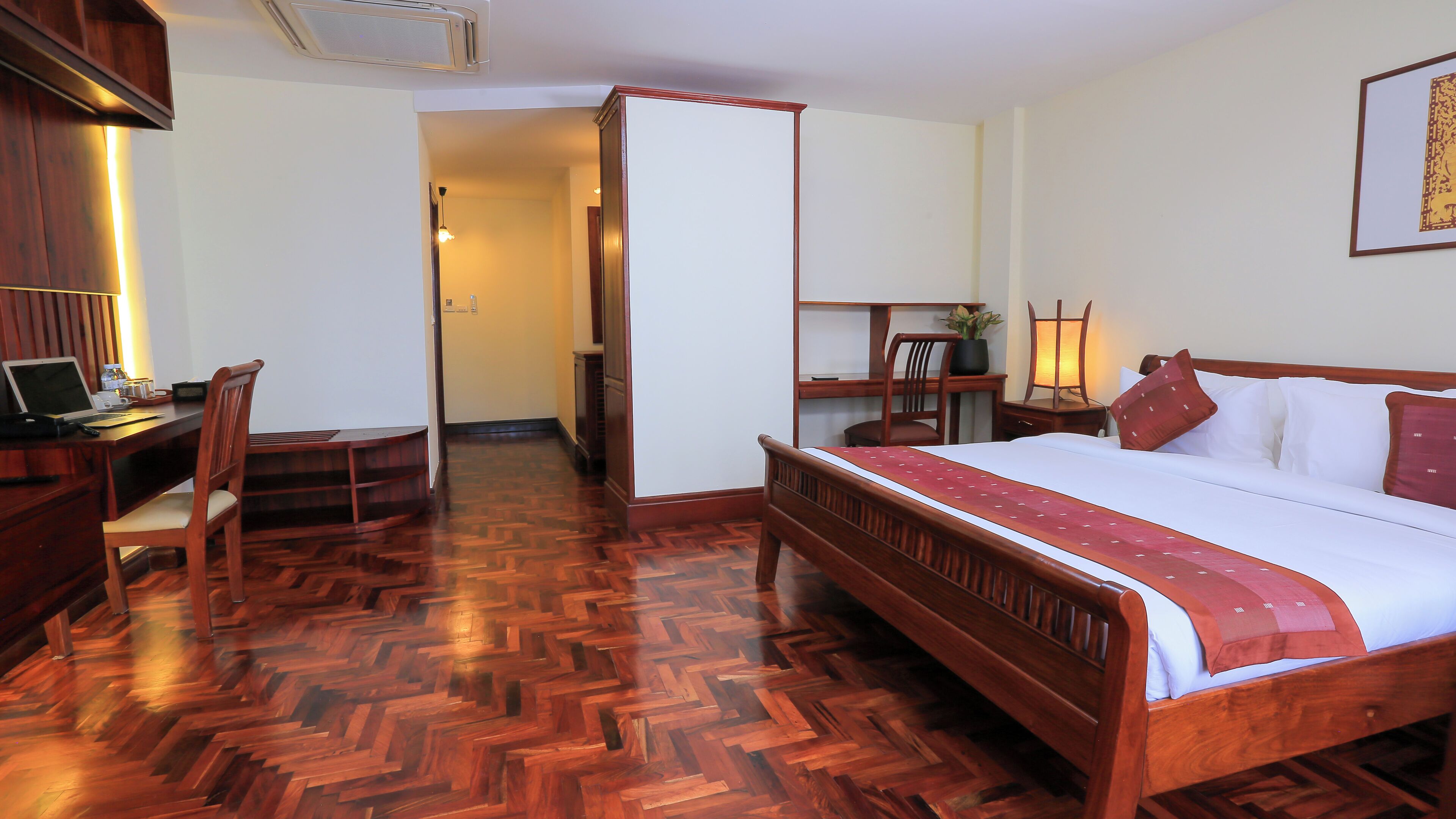 A hotel room with a wooden floor, a bed with white bedding, a desk with a laptop, a chair, and a small table with a plant.