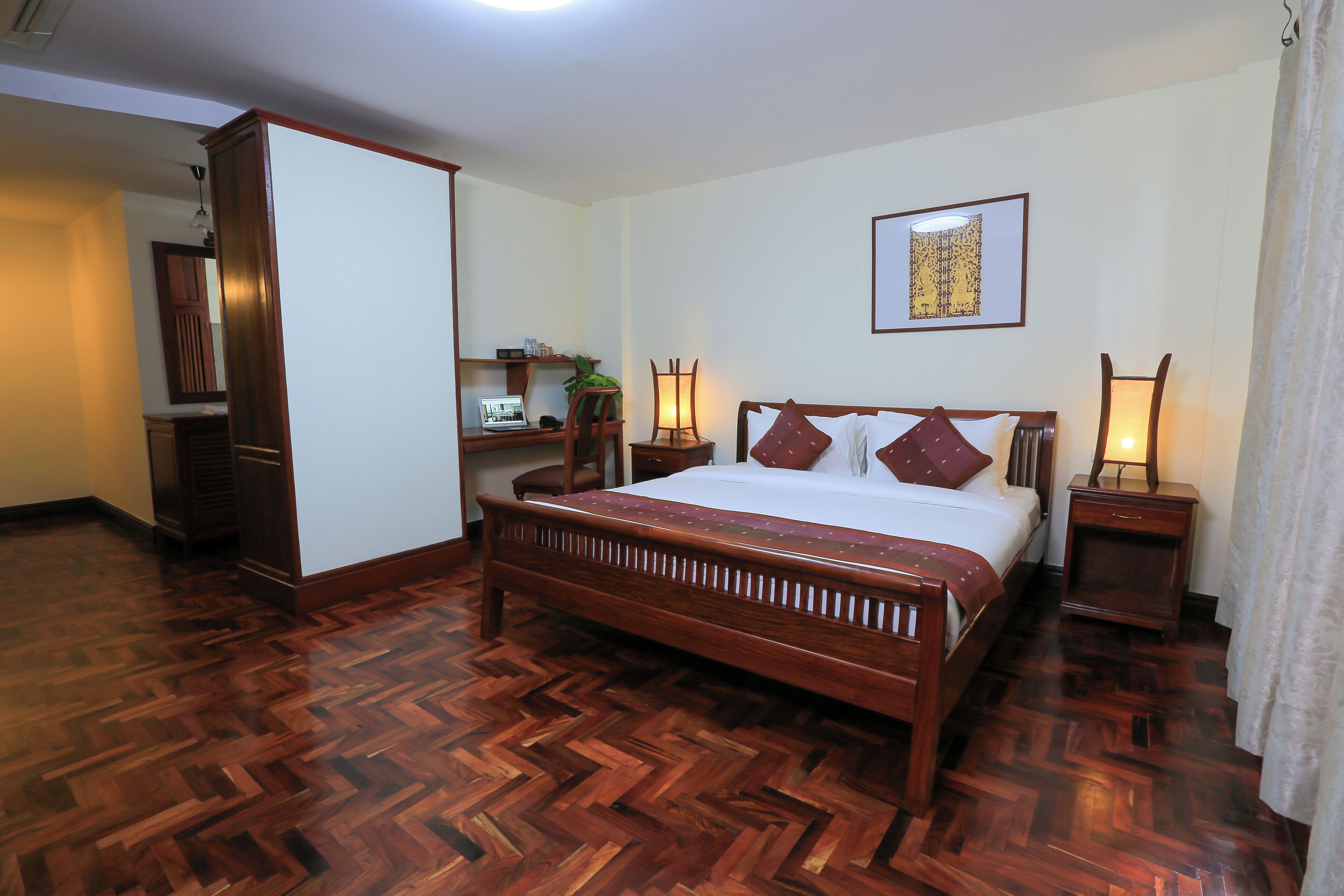 Ein Hotelzimmer mit Holzboden im Fischgrätmuster, einem großen Bett mit Holz-Kopfteil, zwei Nachttischen mit Lampen, einem Schreibtisch mit Stuhl und einem Kleiderschrank.