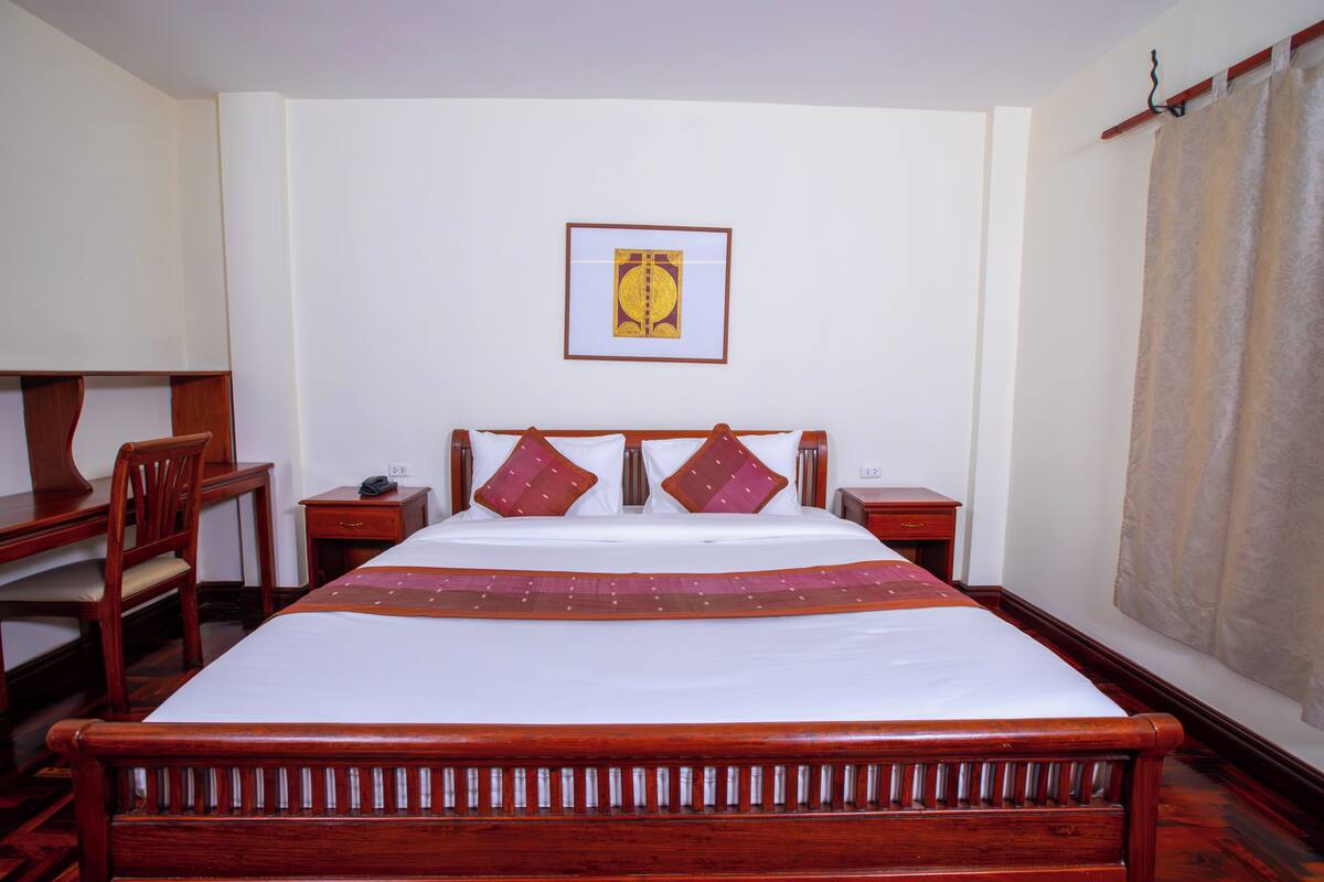 A hotel room with a wooden bed, a desk with a chair, a bedside table, and a framed picture on the wall.