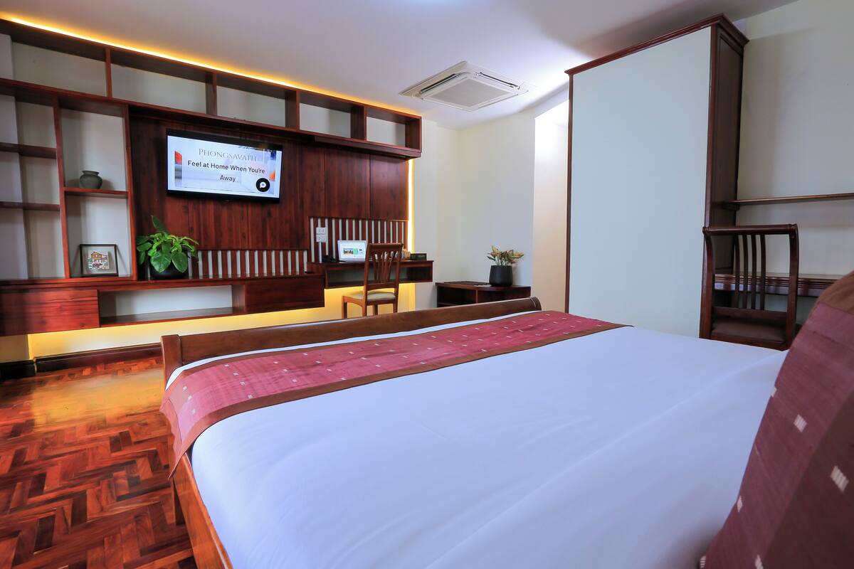 A hotel room with a large bed, wooden headboard, a flat-screen TV mounted on the wall, and a wooden floor.