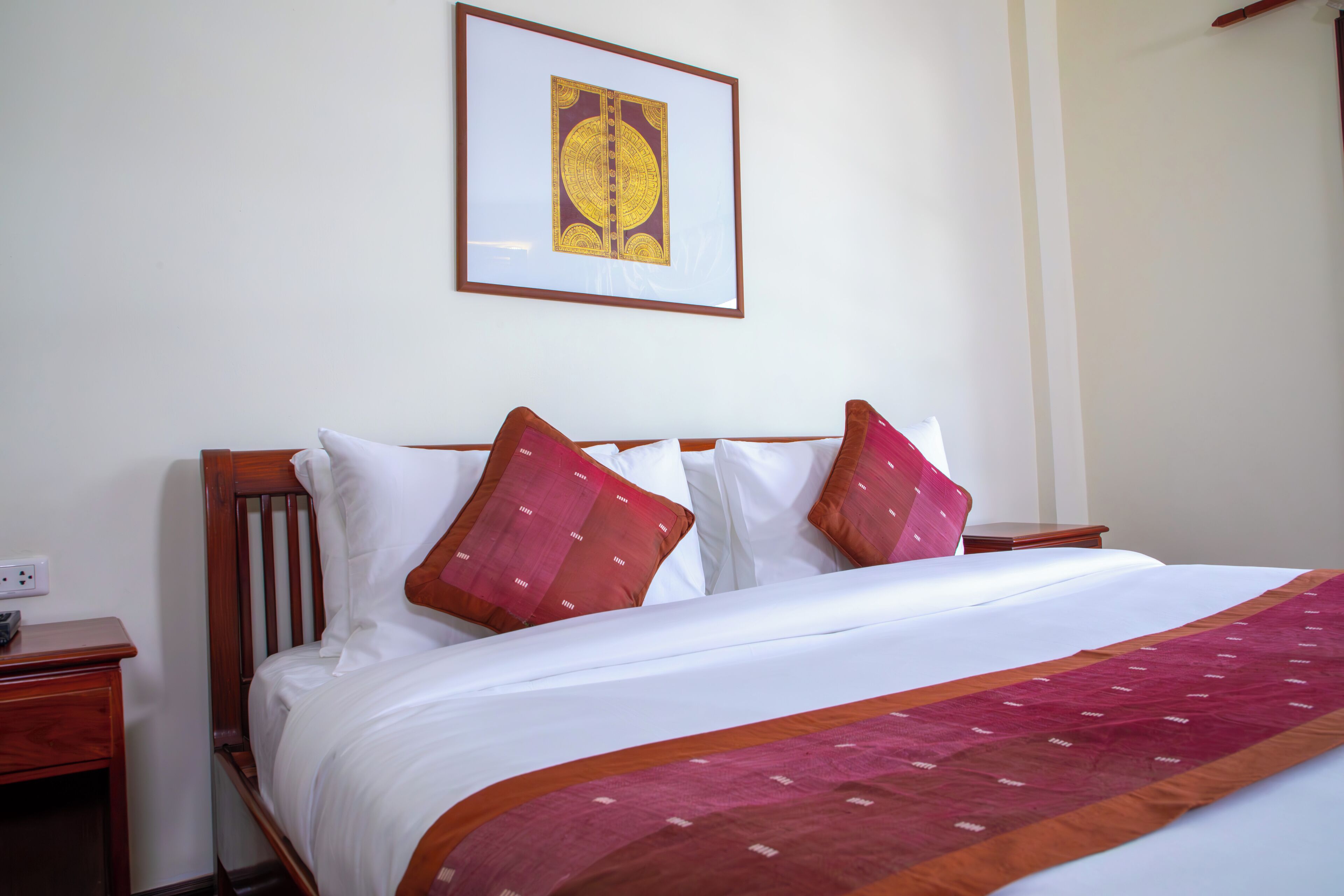 A neatly made bed with white linens and a red and orange patterned throw blanket. There are two wooden nightstands with a small lamp on one. A framed picture hangs on the wall above the bed.