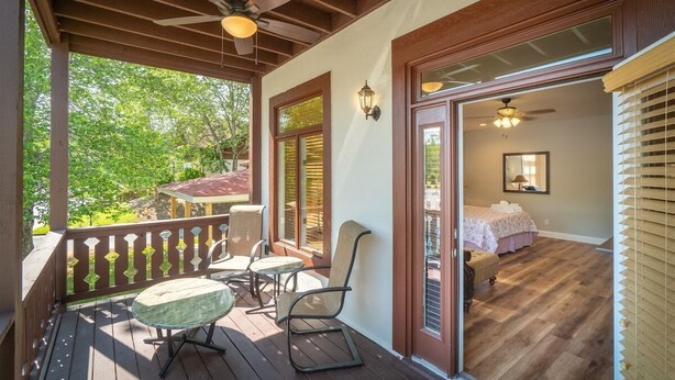 Une terrasse en bois avec des sièges d’extérieur, une chambre avec un lit et une table de chevet, et un ventilateur de plafond.