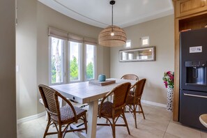 A dining area with a table and chairs, a hanging light fixture, a refrigerator, and a window with blinds.
