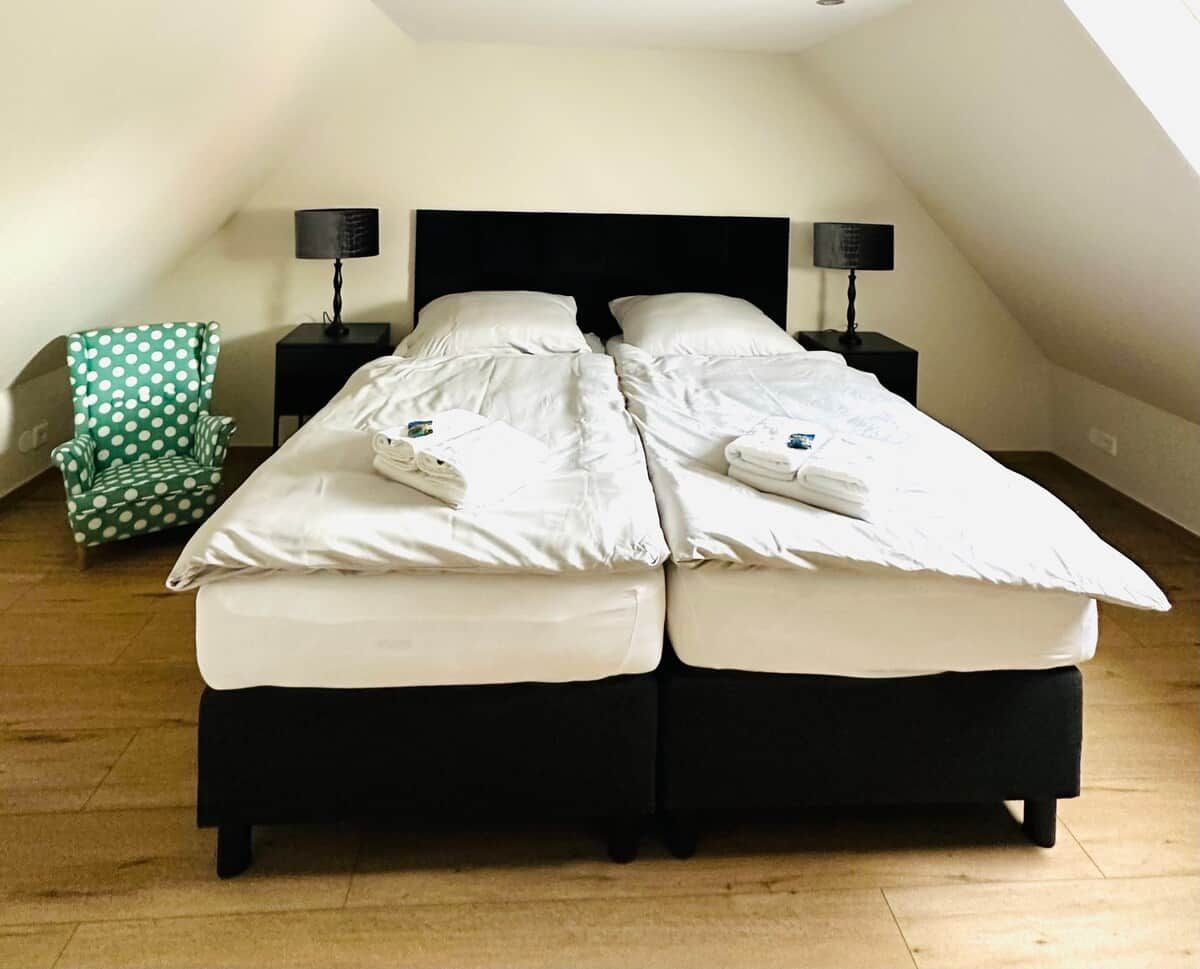 Una cama doble con sábanas blancas, dos mesitas de noche con lámparas, una silla verde con lunares y suelo de madera.
