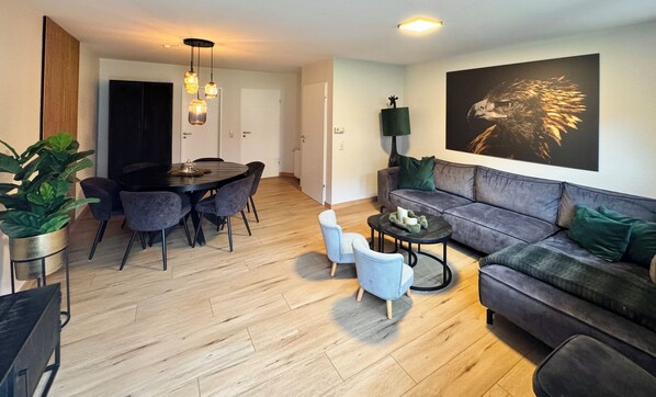 A modern living room with a grey sofa, a round dining table with chairs, a coffee table, and a large painting of an eagle on the wall. - First class house close to the beach, fenced garden 8 adults + children (Varel)