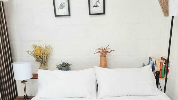 Een netjes opgemaakt bed met witte lakens, een bedlamp, een kleine plant en twee ingelijste foto’s aan de muur.