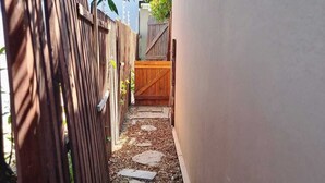 Een smalle steeg met een houten hek aan één kant en een witte muur aan de andere kant. Aan het einde van de steeg bevindt zich een houten poort.