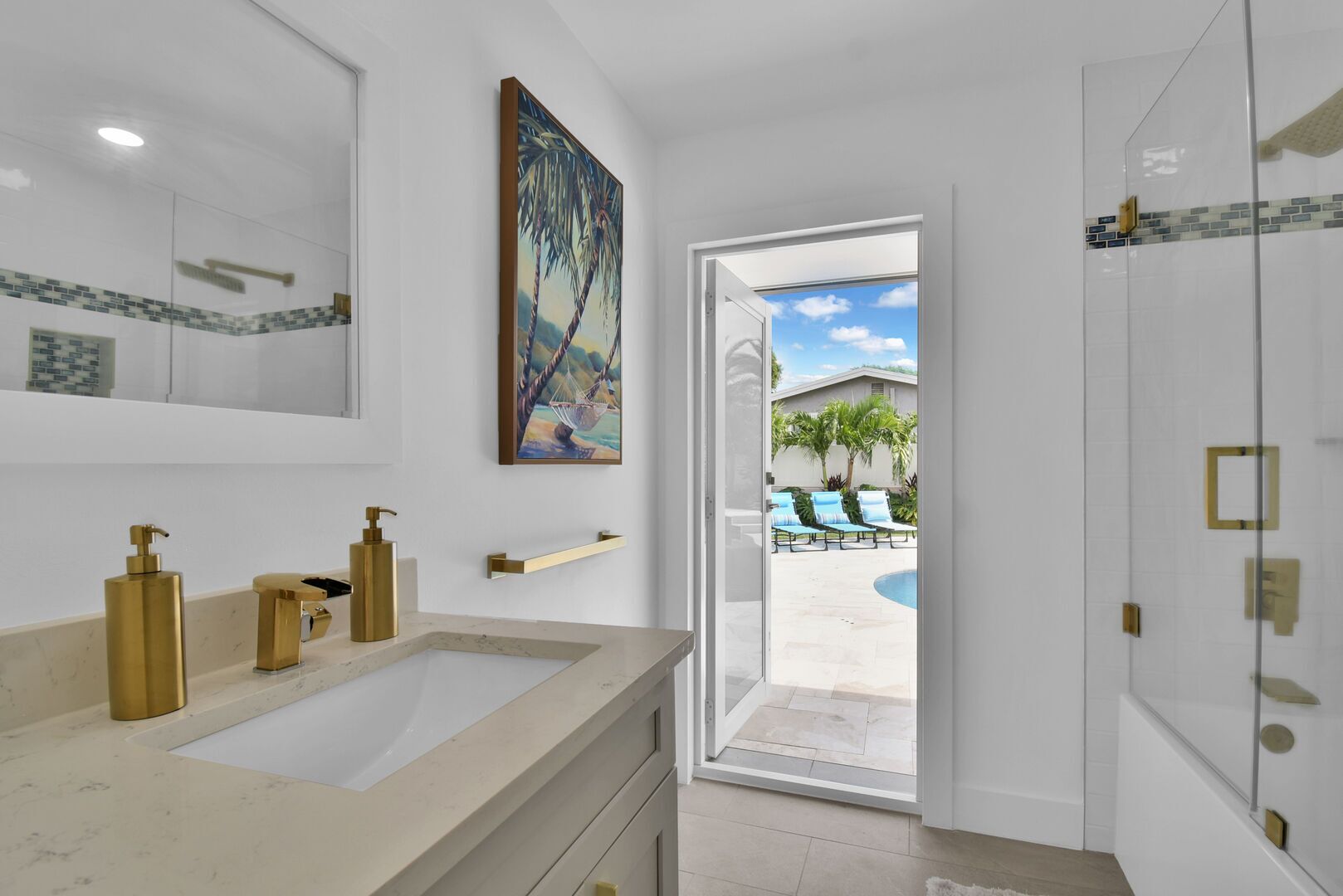 Une salle de bain moderne avec un grand lavabo, des robinetteries dorées et une vue sur une piscine et des chaises longues à l’extérieur.