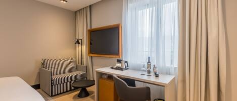 A hotel room with a bed, a sofa, a desk with a computer, a chair, a small cabinet, and a window with curtains.