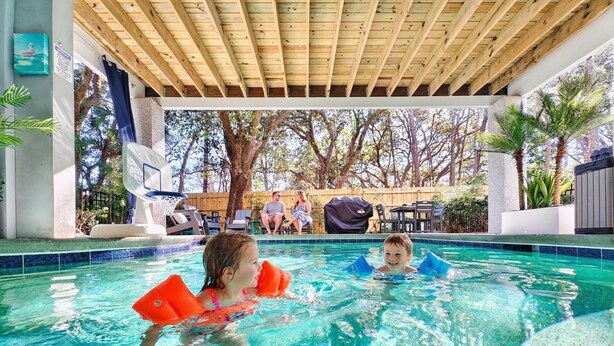 Zona de piscina con techo de madera, agua azul cristalina y vista a un patio trasero con árboles y mobiliario exterior.