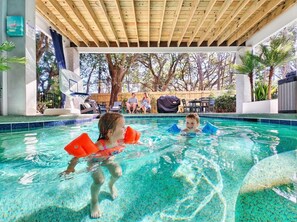 A swimming pool area with a wooden ceiling, clear blue water, and a view of a backyard with trees and outdoor furniture. - Hilton Head 5BR Sleeps 18 • Heated Pool • Walk to Folly Field • Dog Friendly • Elevator (Hilton Head Island)