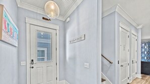 Un couloir avec une porte blanche, un paillasson, une pancarte indiquant « Bienvenue à Beach House », et un escalier menant à une autre pièce.