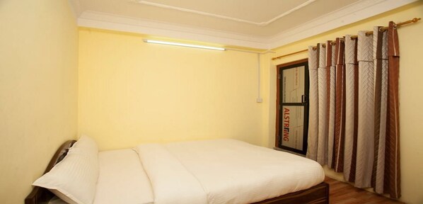 A small, single-bed room with a bed, a nightstand, a wall-mounted light, and a window with curtains. - MeroStay 261 Hotel Wall (Kathmandu)
