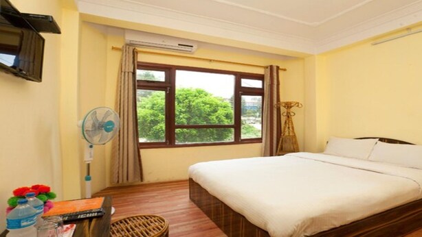 A hotel room with a bed, a window with curtains, a standing fan, and a small table with a water bottle and a remote control.