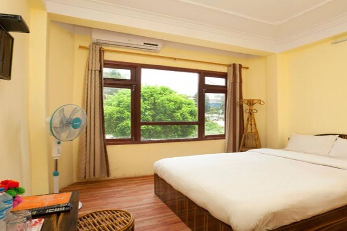 A hotel room with a bed, a window with curtains, a standing fan, and a small table with a water bottle and a remote control.