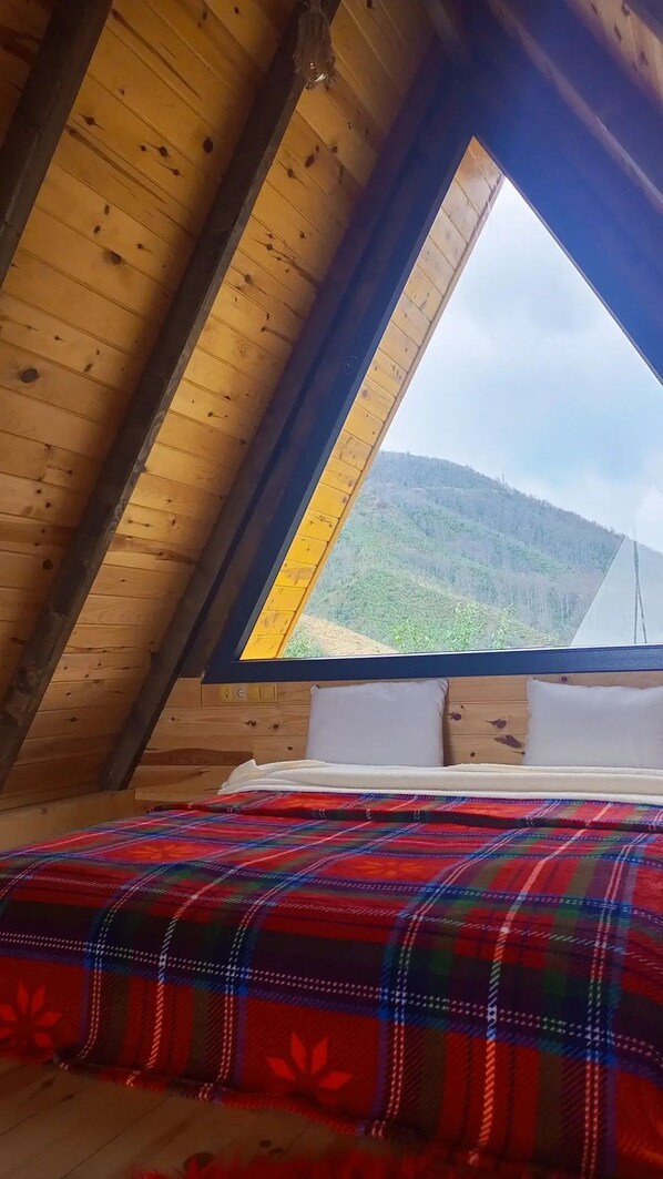 A bedroom with a sloped ceiling, a wooden bed frame, a colorful plaid bedspread, and a window offering a view of a hilly landscape. - Pamira Bungalow (Surmene)