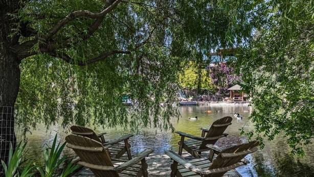 Een houten terras met stoelen, uitkijkend op kalm water, omgeven door weelderige begroeiing en een prieeltje op de achtergrond.