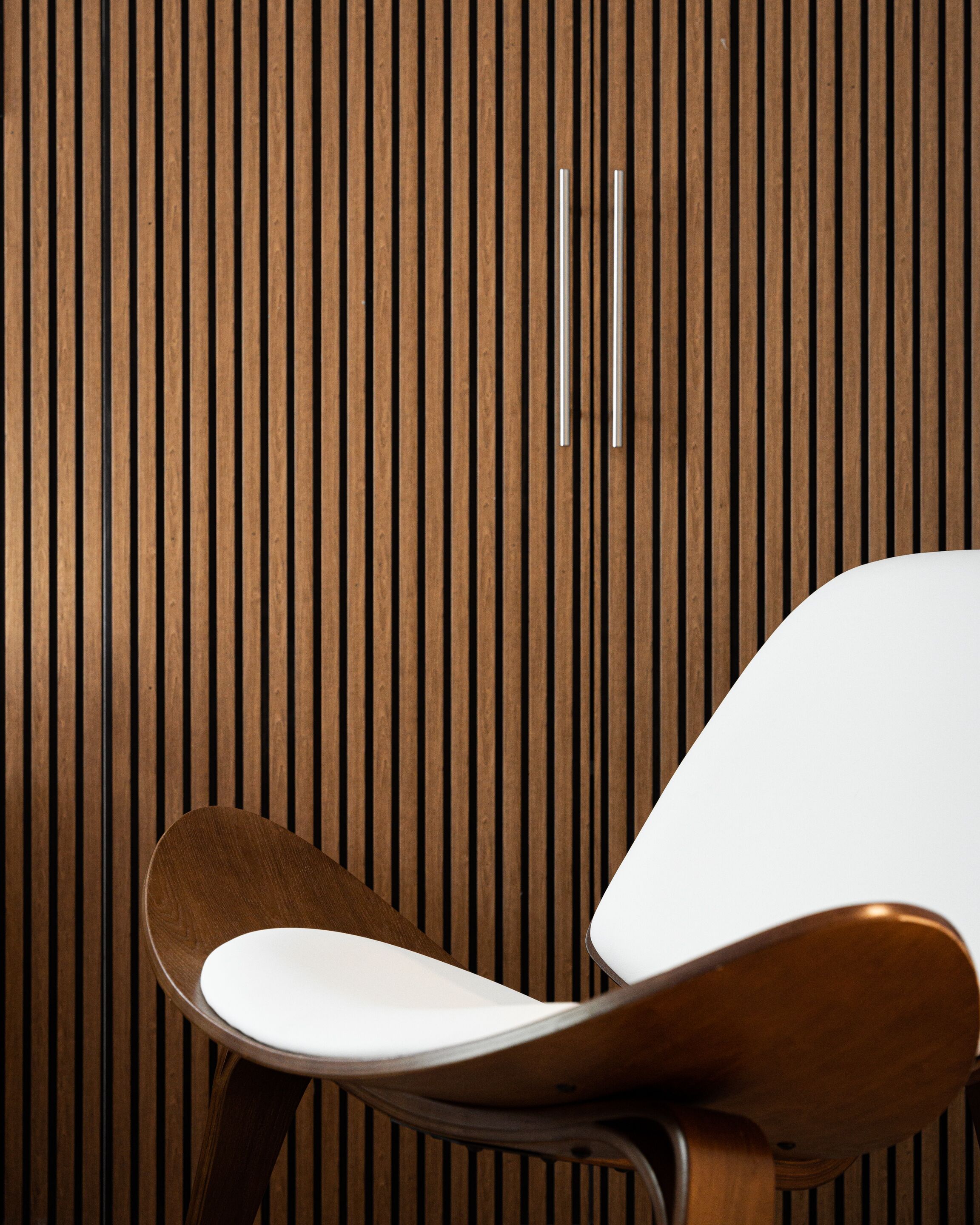 A modern chair with a wooden frame and white seat, positioned in front of a wooden slat wall with two metallic handles.