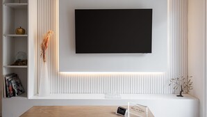 Una sala de estar moderna con televisor montado, estanterías blancas con objetos decorativos y una mesa de madera con un cartel.