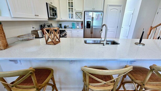 A kitchen with white cabinets, a stainless steel refrigerator, a sink, and wicker chairs. - Coastal Club * 19117 Twin House w 4BR, 3.5 Baths, Sleeps 11 (Lewes)