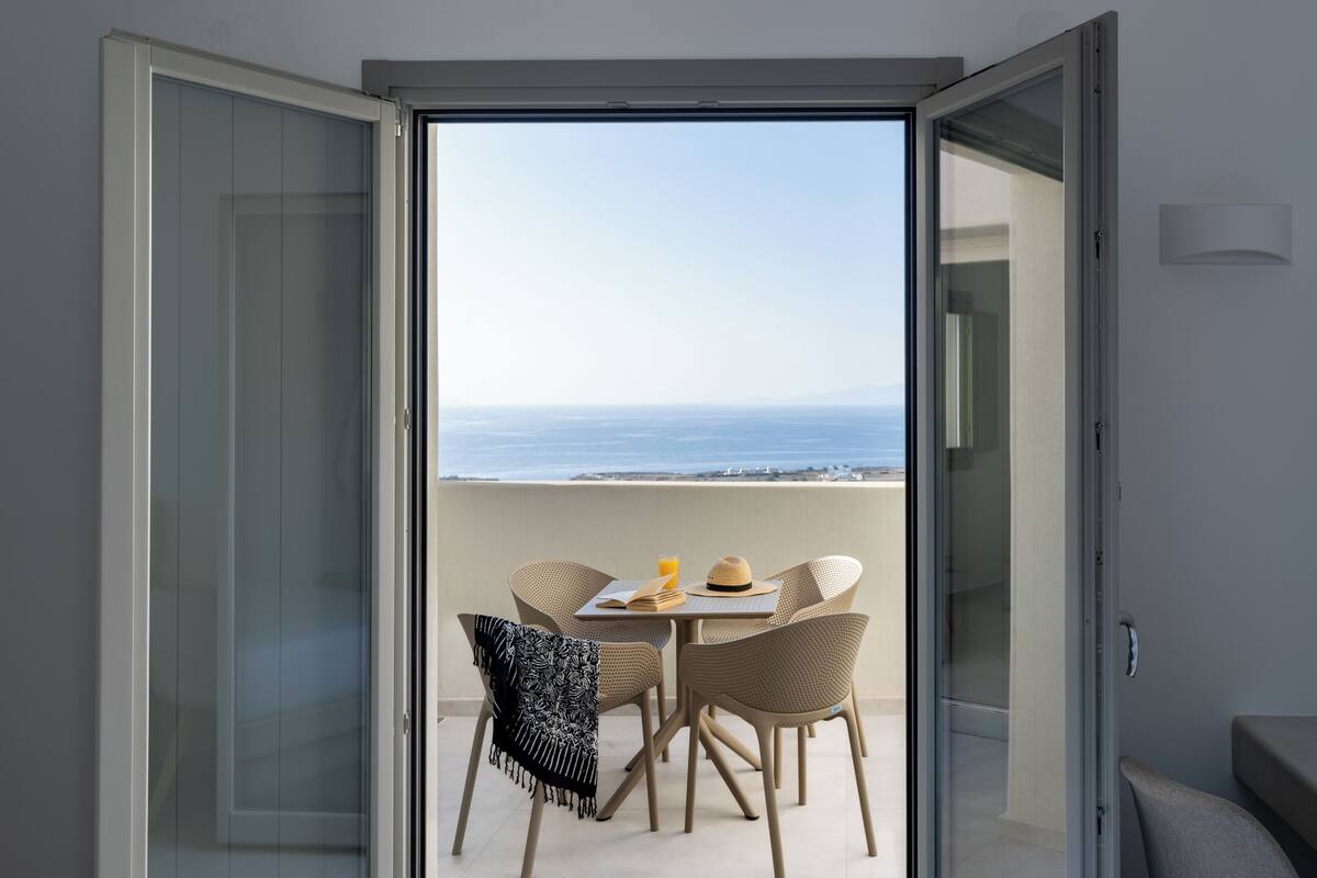 Eine offene Schiebetür führt zu einem Balkon mit Tisch, Stühlen und Blick auf das Meer.