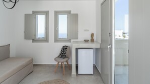 Un espace de vie compact comprenant un canapé, un fauteuil, une petite table et un réfrigérateur. Il y a deux fenêtres avec des volets ouverts, et on aperçoit une porte-fenêtre donnant sur le balcon.