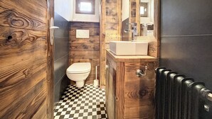 A bathroom with a wooden wall design, a white toilet, a sink with a mirror, and a black radiator. - Living room with eating area and a fireplace, a kitchen with dishwasher and Nespresso Coffeemachine, (Verbier)