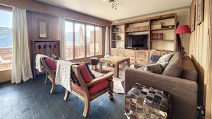 A cozy living room with a fireplace, wooden furniture, a bookshelf, and large windows offering a mountain view. - Living room with eating area and a fireplace, a kitchen with dishwasher and Nespresso Coffeemachine, (Verbier)