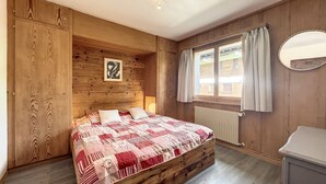 A wooden-paneled bedroom with a bed, a window with curtains, a radiator, and a mirror on the wall. - Living room with eating area and a fireplace, a kitchen with dishwasher and Nespresso Coffeemachine, (Verbier)