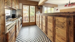 A kitchen with wooden cabinets, a black and white checkered floor, and a view through a glass door to an outdoor deck. - Living room with eating area and a fireplace, a kitchen with dishwasher and Nespresso Coffeemachine, (Verbier)