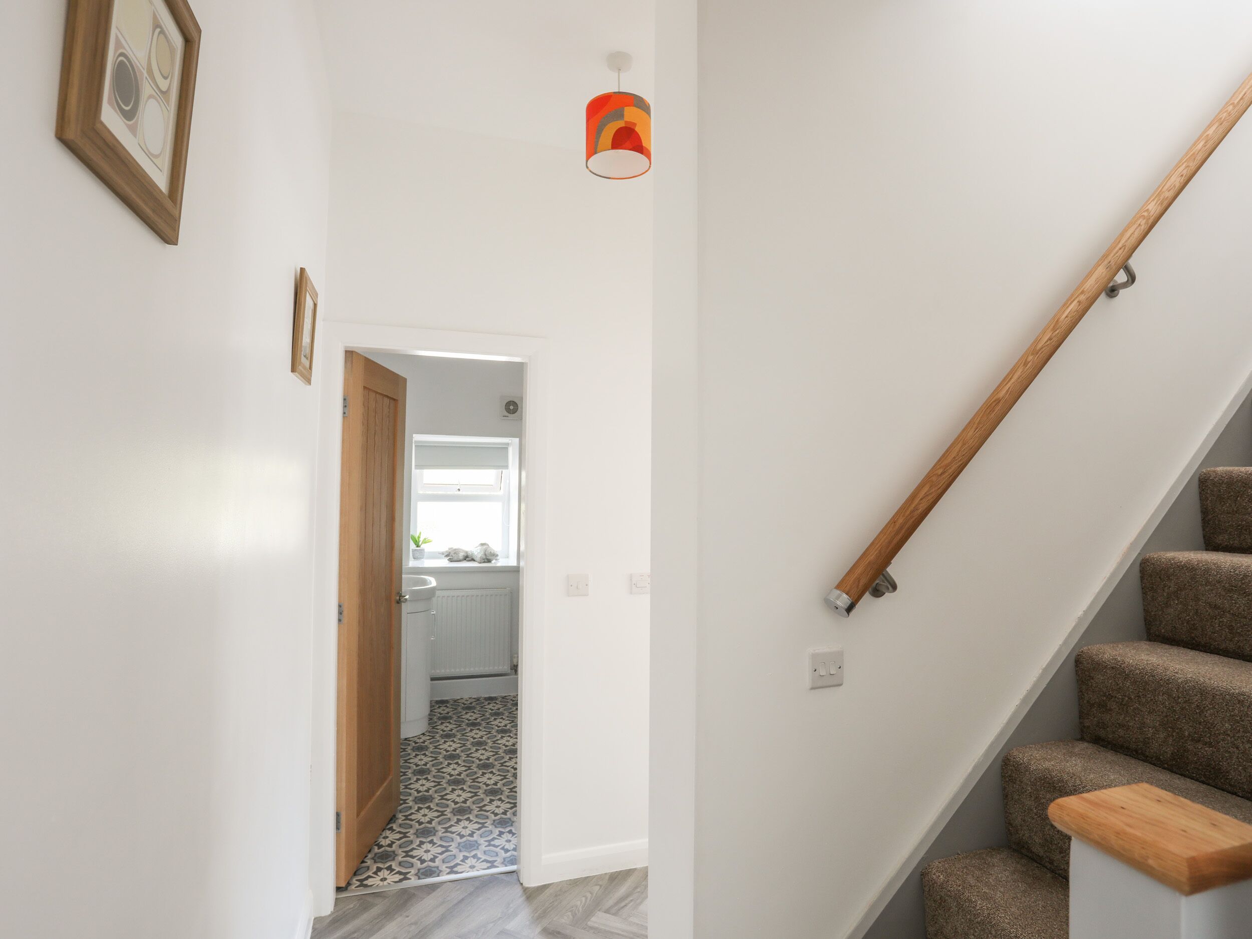 Un pasillo con una escalera de madera, una puerta que da al baño y un pequeño ventanal con una planta.
